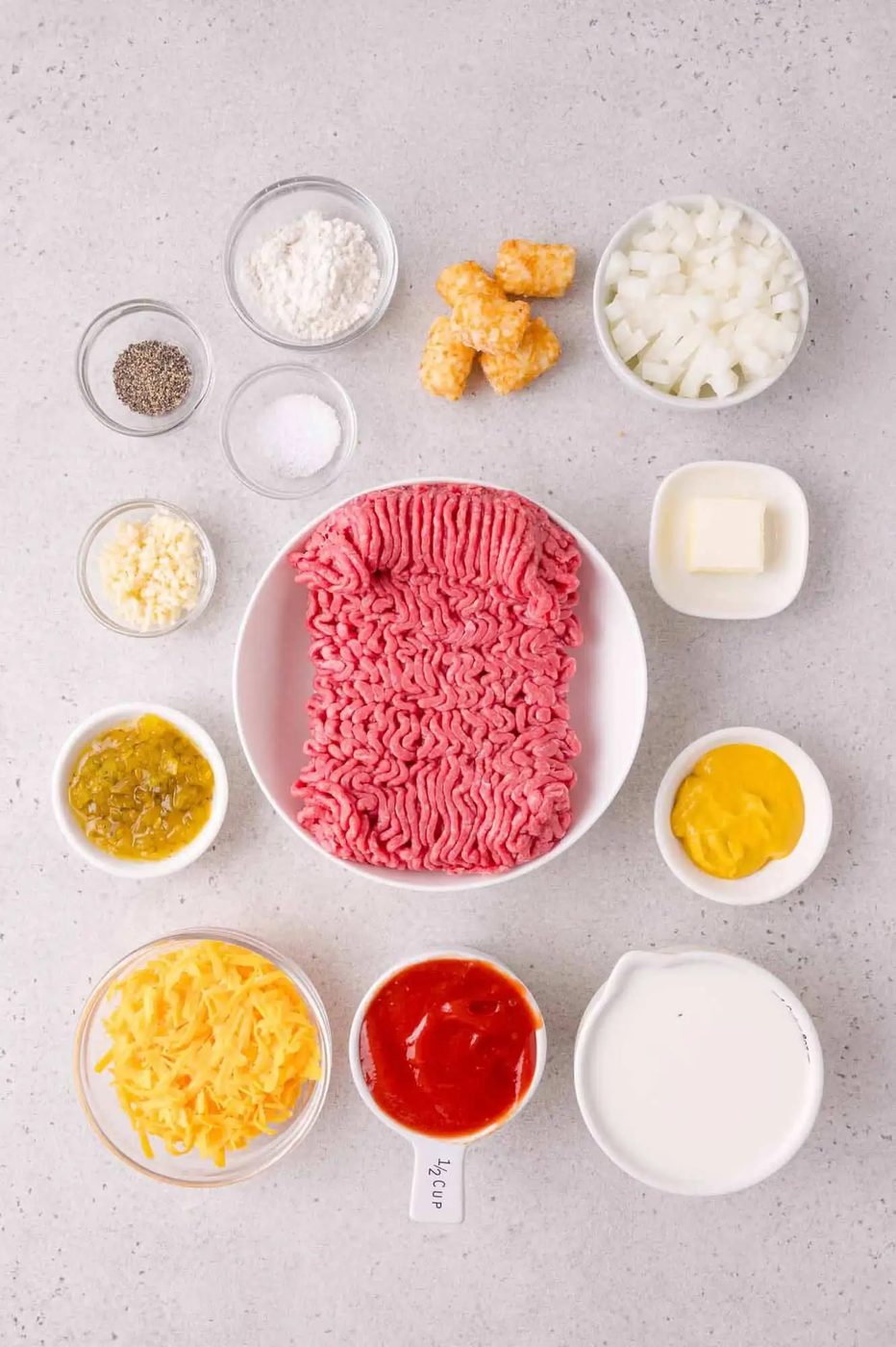 Ground beef, shredded cheese, diced onions, tater tots, milk, ketchup, mustard, relish, butter, flour, salt, pepper, and garlic arranged in bowls on a gray countertop.