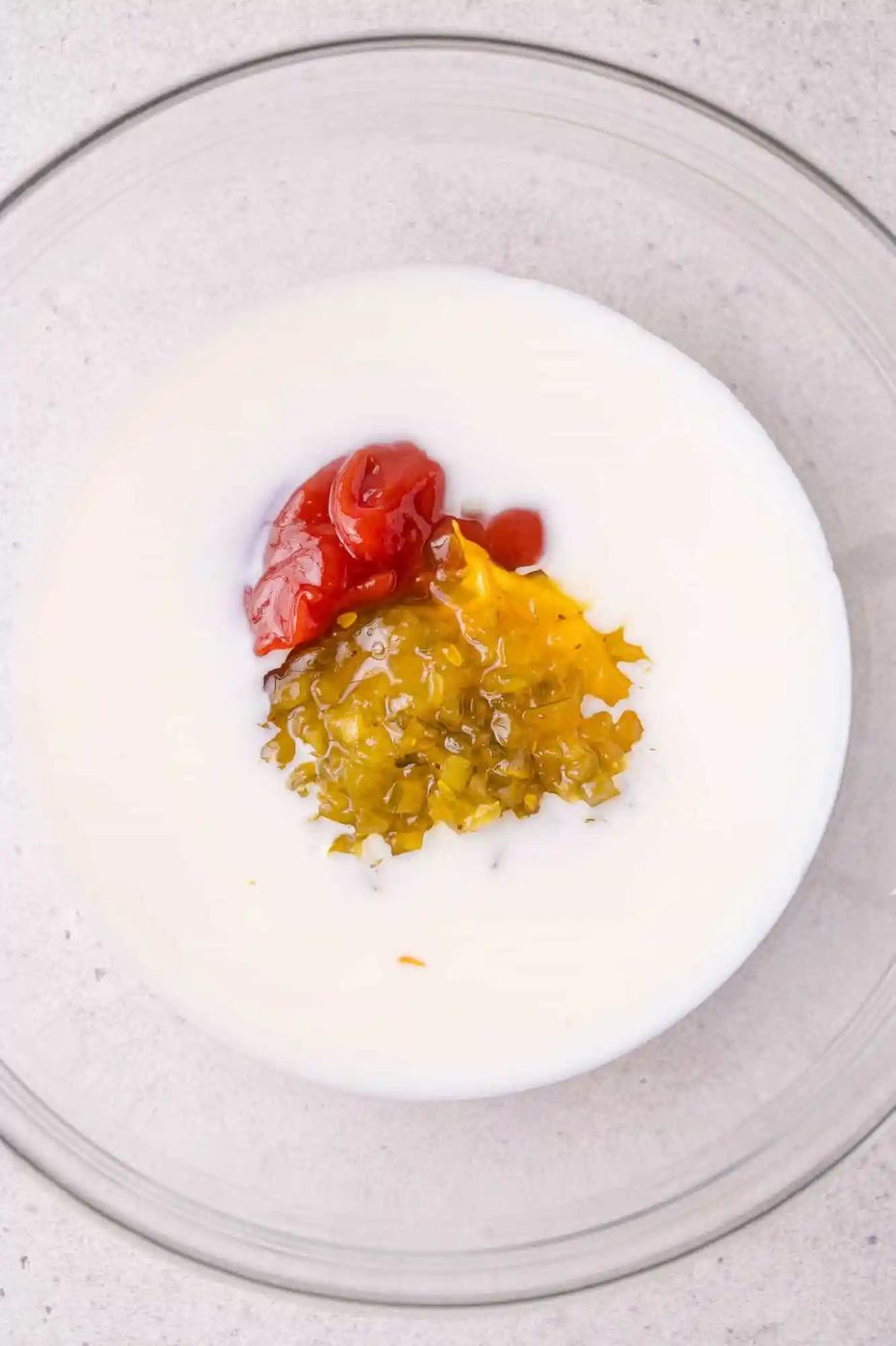 A glass bowl containing milk, ketchup, yellow mustard, and relish on a light surface.