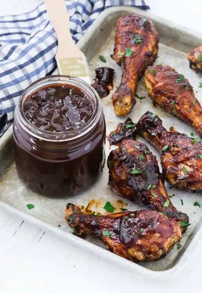A sheet pan with grilled chicken drumsticks, and a jar of from-scratch barbecue sauce. A blue and white checkered linen is also pictured.