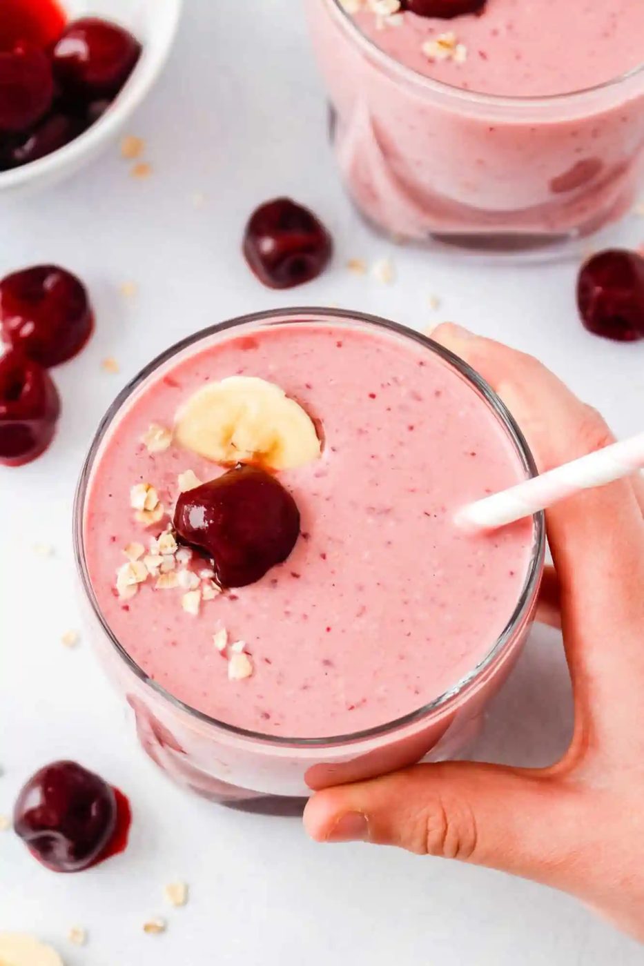 A hand holds a glass of pink cherry smoothie topped with a banana slice, cherry, and oats, with a straw inserted. Other cherries are scattered on the white surface.