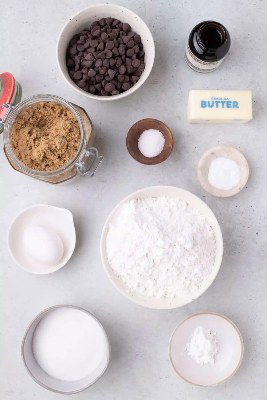 Ingredients for baking: chocolate chips, vanilla extract, butter, brown sugar, salt, baking soda, flour, egg, white sugar, and baking powder are arranged on a countertop.