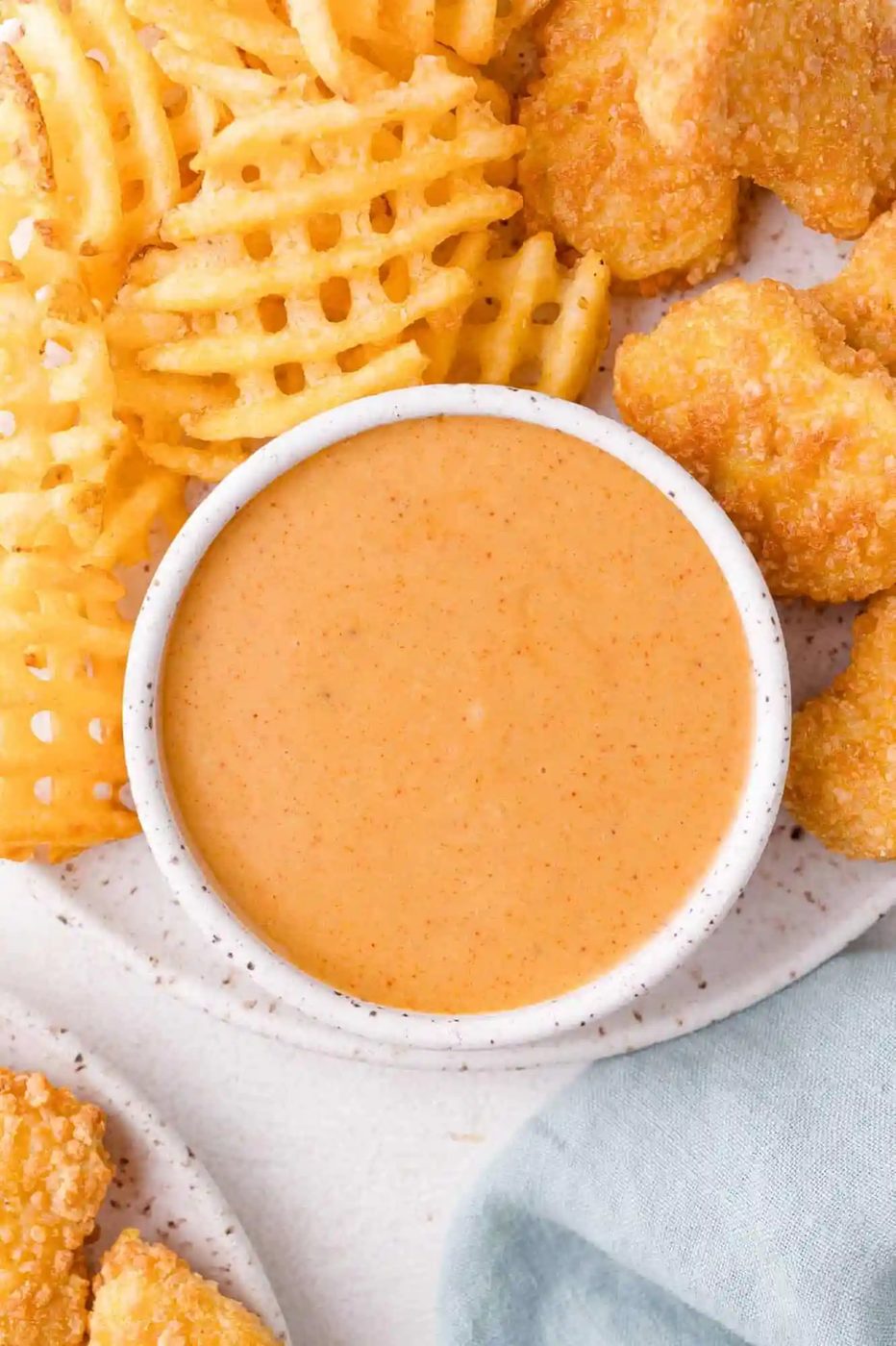 A bowl of creamy dipping sauce is surrounded by waffle fries and breaded chicken nuggets on a plate.