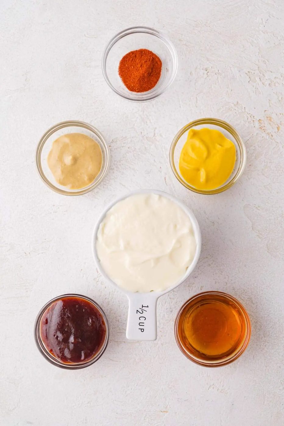 Six small containers with various condiments and spices, including mayonnaise, mustard, honey, barbecue sauce, Dijon mustard, and paprika, arranged on a light surface.