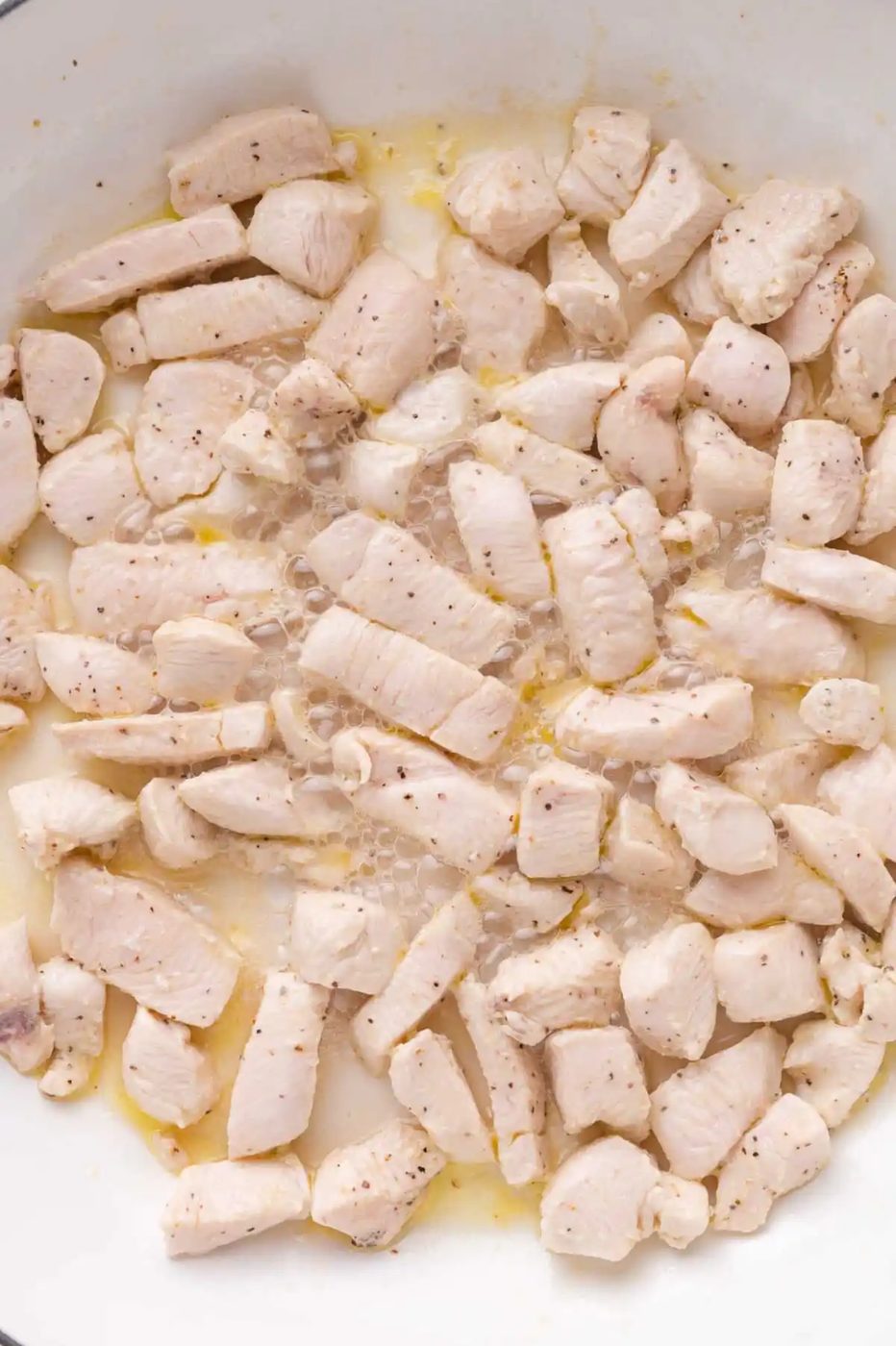 Cubed pieces of chicken breast being cooked in a pan, lightly seasoned, with visible moisture and slight browning.