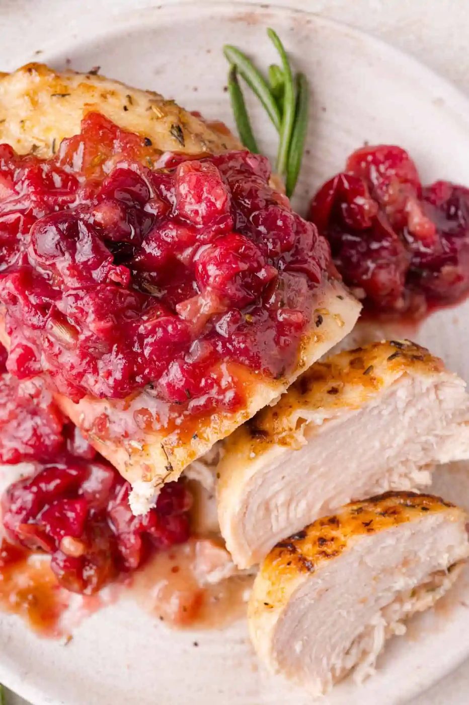 Cooked chicken breast topped with a chunky cranberry sauce, garnished with fresh rosemary on a white plate.