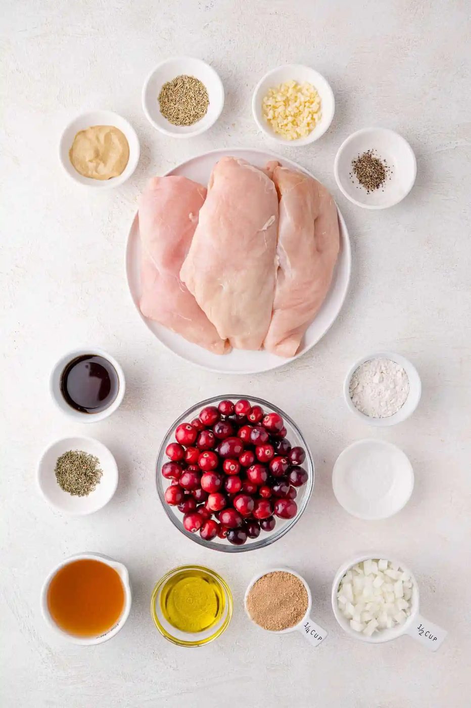 Top view of ingredients for a cranberry chicken dish on a light surface, including raw chicken breasts, cranberries, herbs, spices, sauces, and diced onions each in separate bowls.