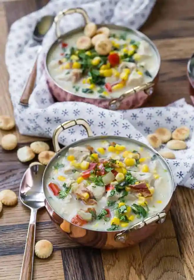 Corn chowder in a copper bowl, two bowls visible, with a blue and white linen and oyster crackers also pictured.