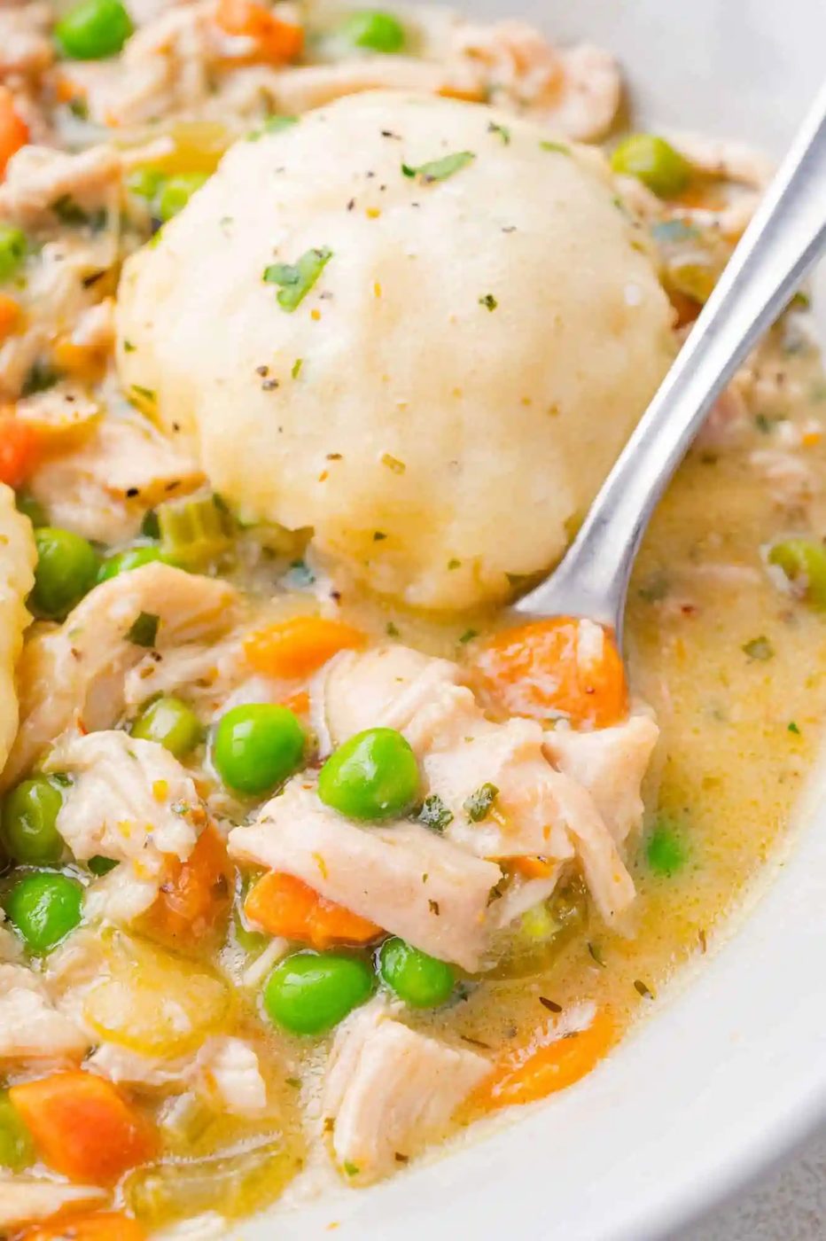 A bowl of chicken dumpling soup with peas and carrots, topped with a dumpling. A spoon is in the bowl.
