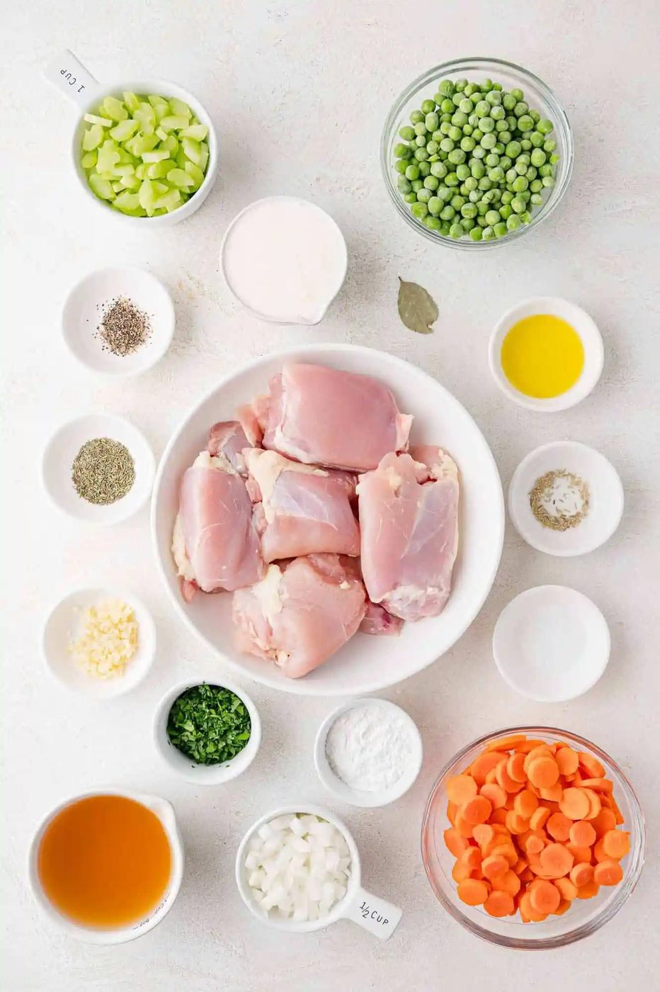 Ingredients arranged on a white surface: raw chicken thighs, sliced carrots, peas, chopped celery, garlic, onion, herbs, seasonings, broth, cream, olive oil, and flour.