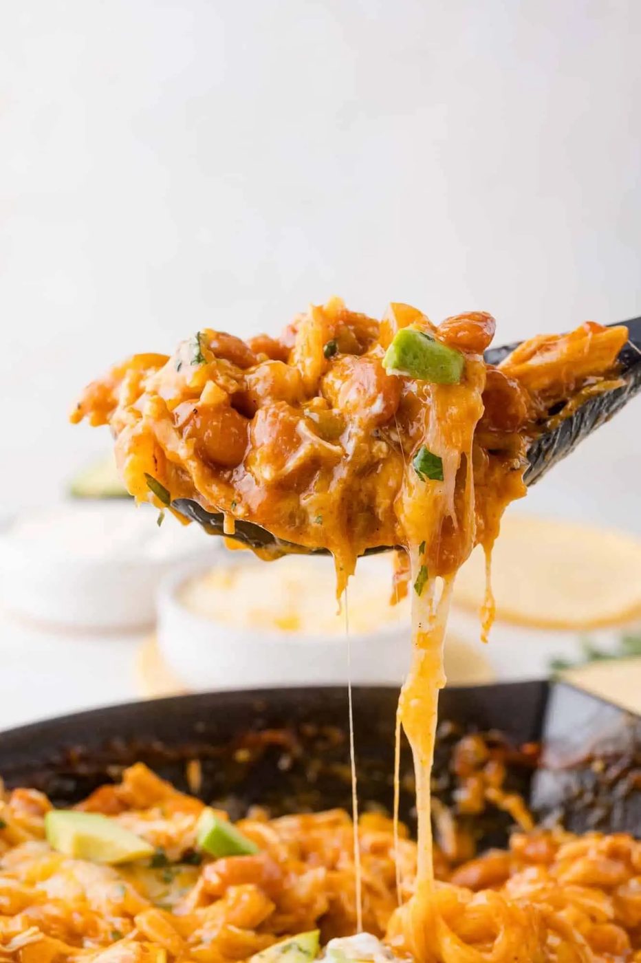 A close-up of a spoon lifting a cheesy, bean and pasta skillet dish, with melted cheese stretching from the pan.