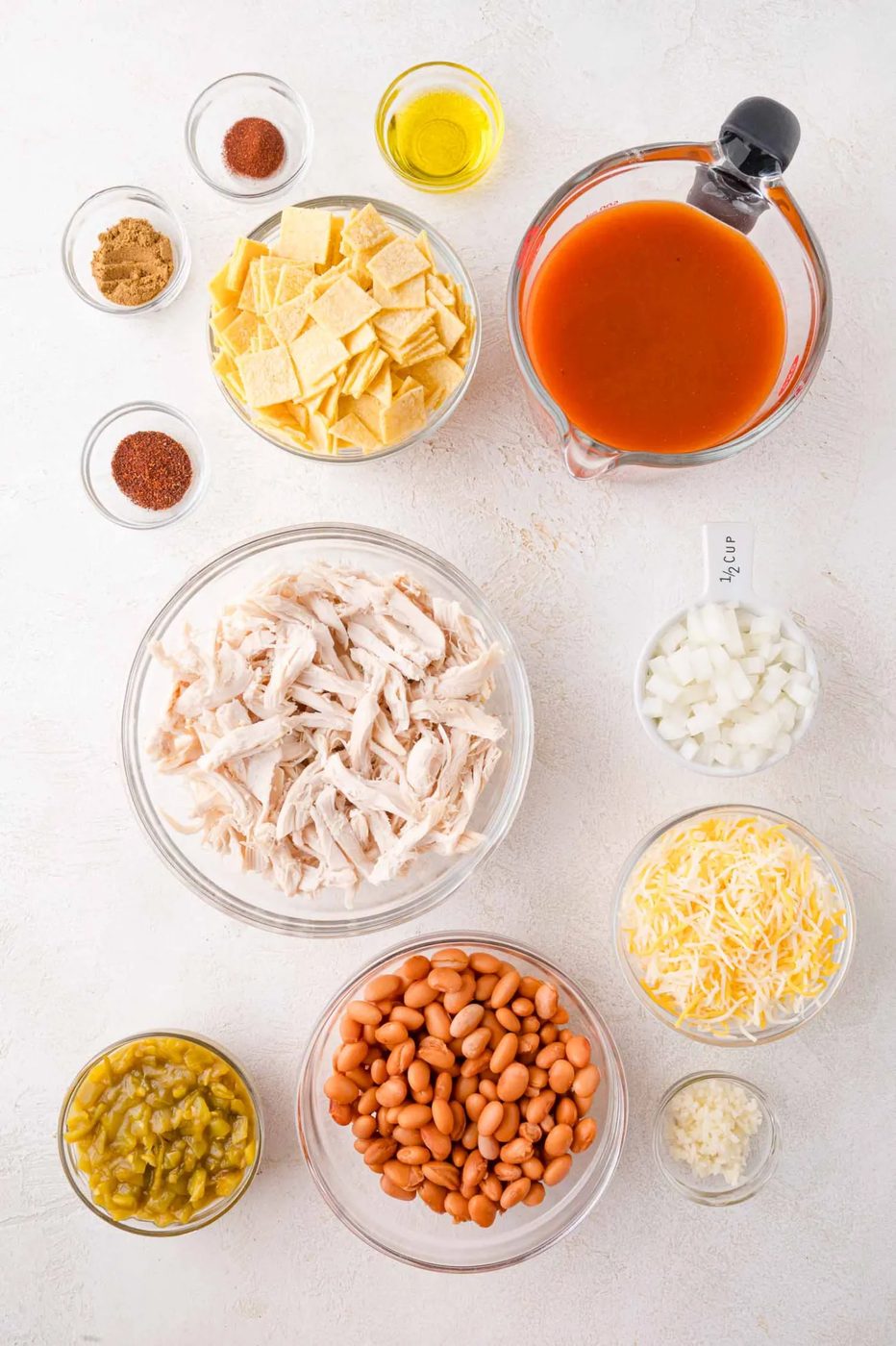 Various ingredients are arranged in bowls on a white surface, including shredded chicken, pinto beans, cheese, chopped onions, enchilada sauce, tortilla pieces, and seasonings.