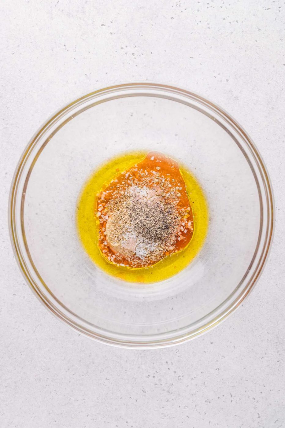 A clear glass bowl containing olive oil, honey, and seasonings on a light countertop.