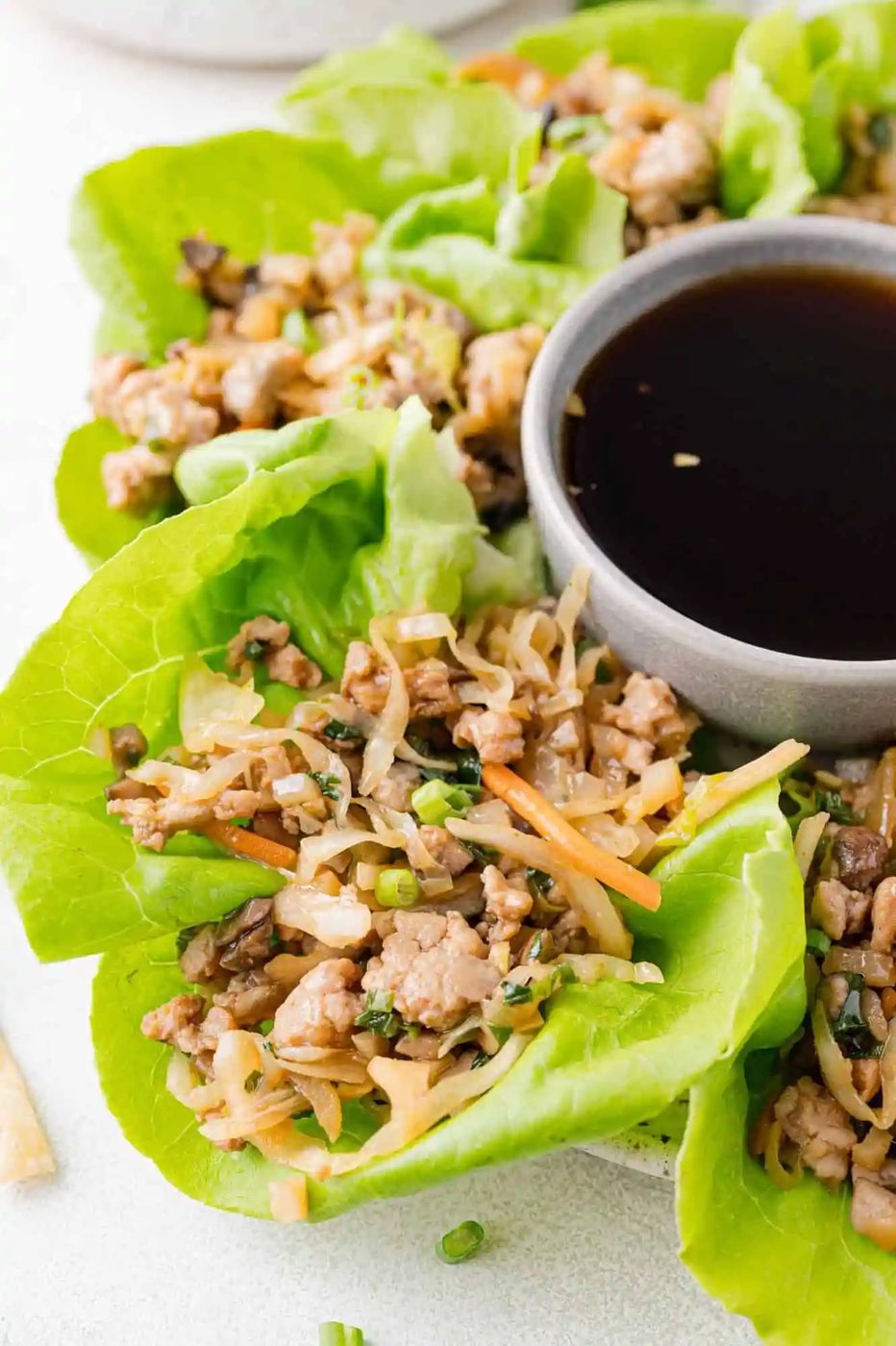 Fresh lettuce wraps filled with minced chicken, vegetables, and herbs are arranged around a small bowl of dipping sauce on a white background.