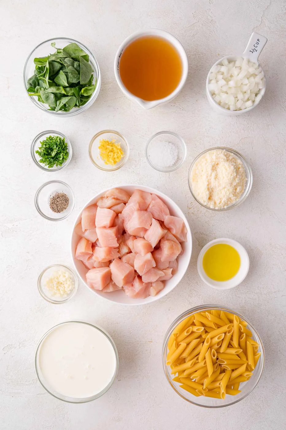 Assorted ingredients on a white surface, including chopped chicken, penne pasta, spinach, broth, cream, onions, cheese, oil, garlic, herbs, and seasonings in separate bowls.