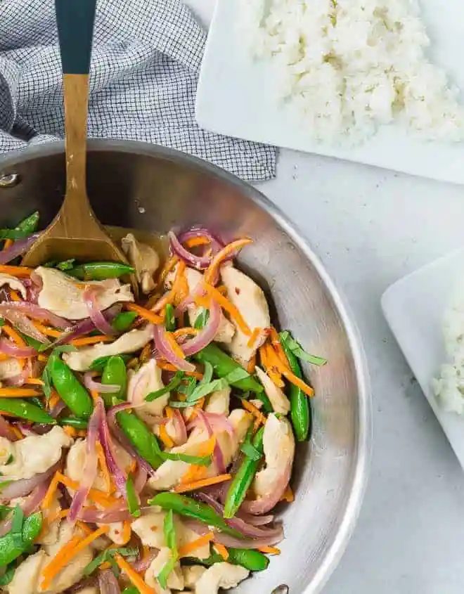 Overhead view of a wok filled with white rice, chicken, sugar snap peas, carrots, and red onions in stir fry form. It is garnished with fresh basil ribbons.