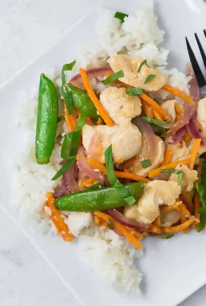Overhead view of a white plate filled with white rice, chicken, sugar snap peas, carrots, and red onions in stir fry form. It is garnished with fresh basil ribbons.