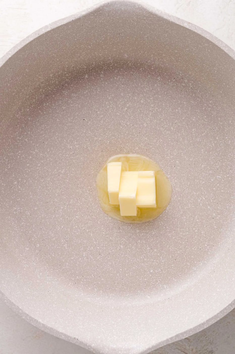 A few pieces of butter melting in the center of a light-colored, empty skillet.