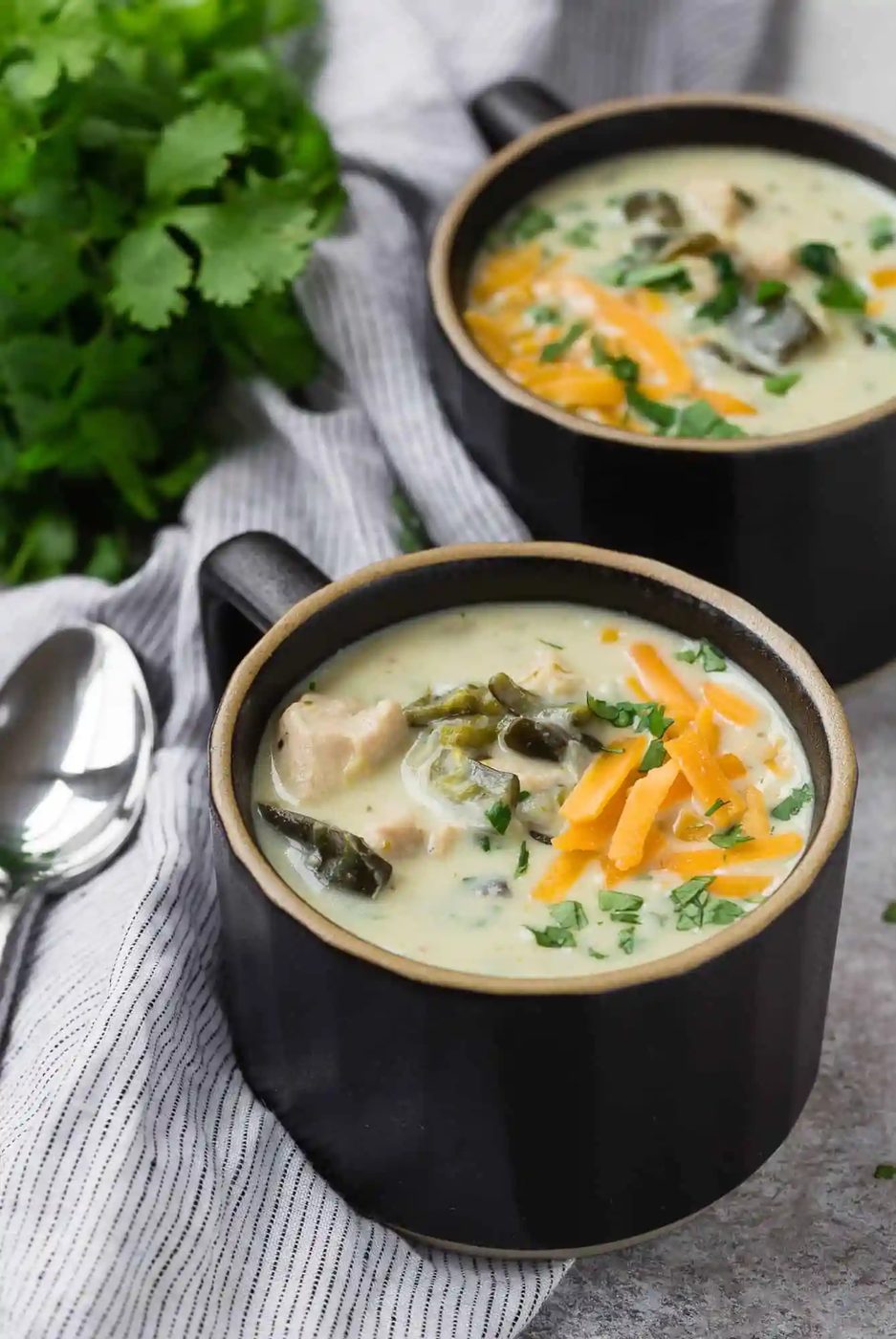 Two mug-style bowls of chicken chile relleno soup, topped with cheddar.
