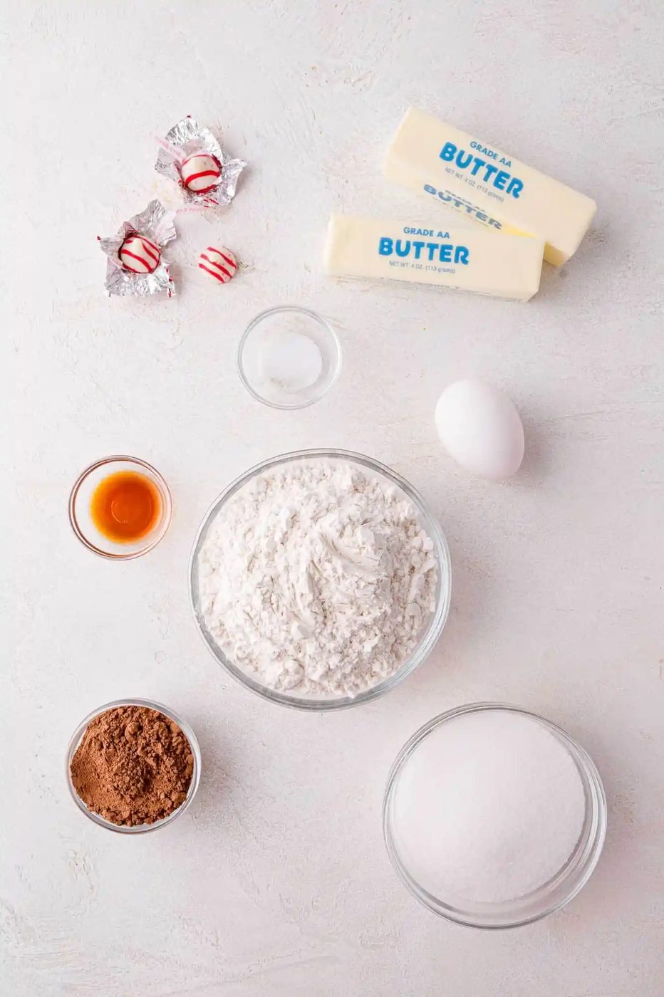 Baking ingredients on a countertop include flour, sugar, butter, cocoa powder, an egg, vanilla extract, salt, and peppermint candies.