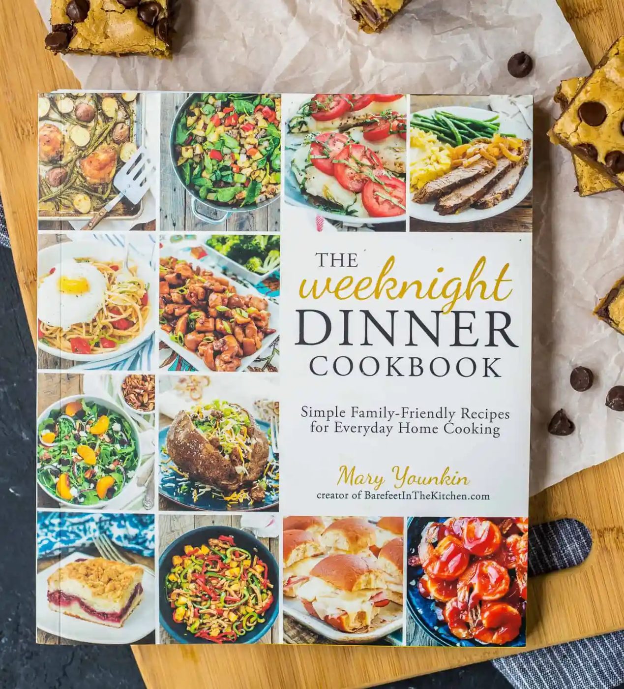The Weeknight Dinner Cookbook by Mary Younkin is on a wooden surface beside chocolate chip bars and scattered chocolate chips.