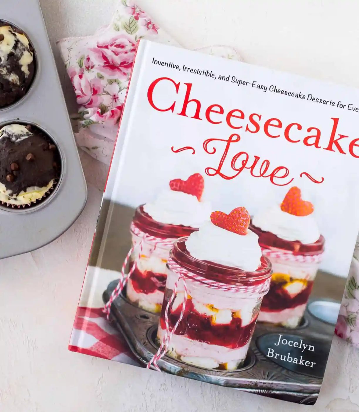 A cookbook titled Cheesecake Love by Jocelyn Brubaker is on a floral cloth next to a muffin tin with two baked muffins.