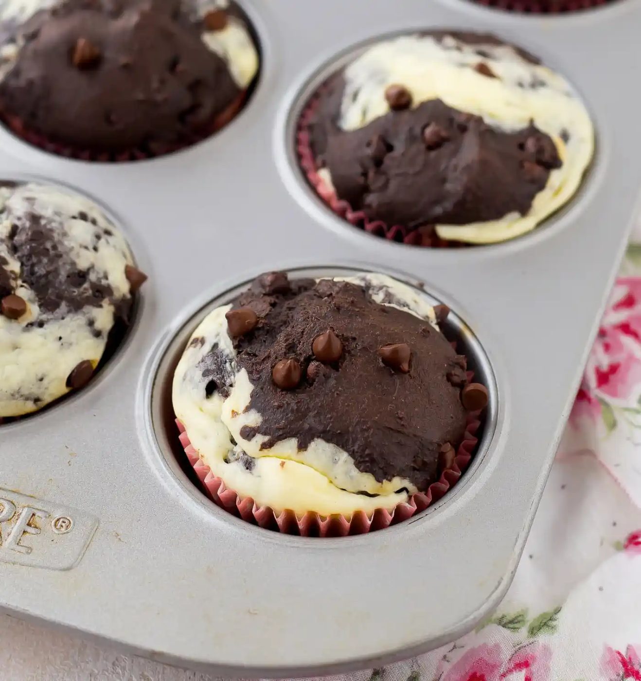 A muffin tin with marbled chocolate and cream cheese muffins, each topped with chocolate chips, on a floral-patterned cloth.