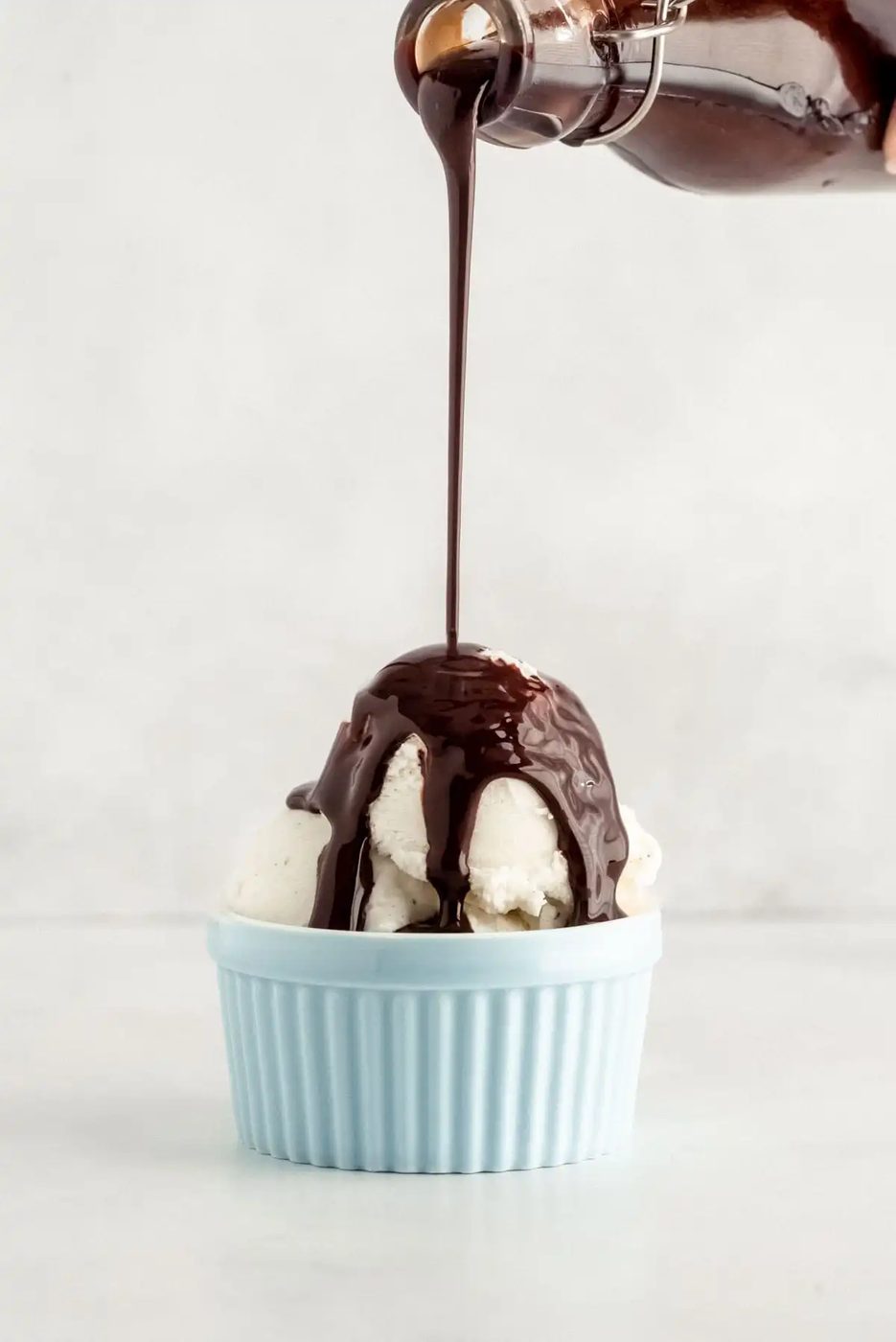 Chocolate syrup being poured onto vanilla ice cream in a light blue bowl against a plain background.