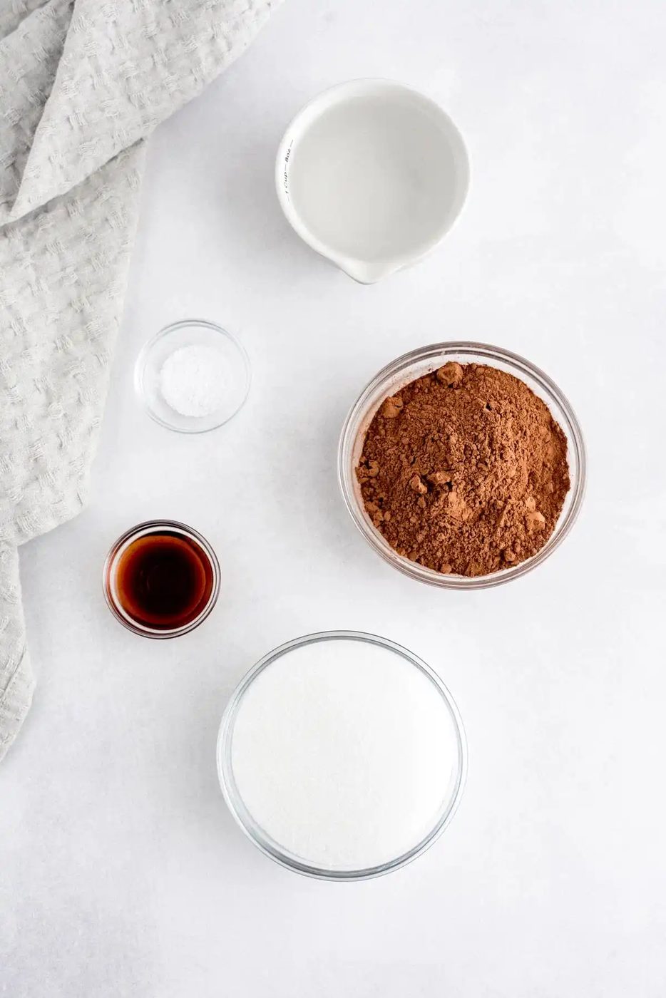 Bowls containing cocoa powder, sugar, vanilla extract, salt, and water arranged on a white surface next to a textured gray cloth.