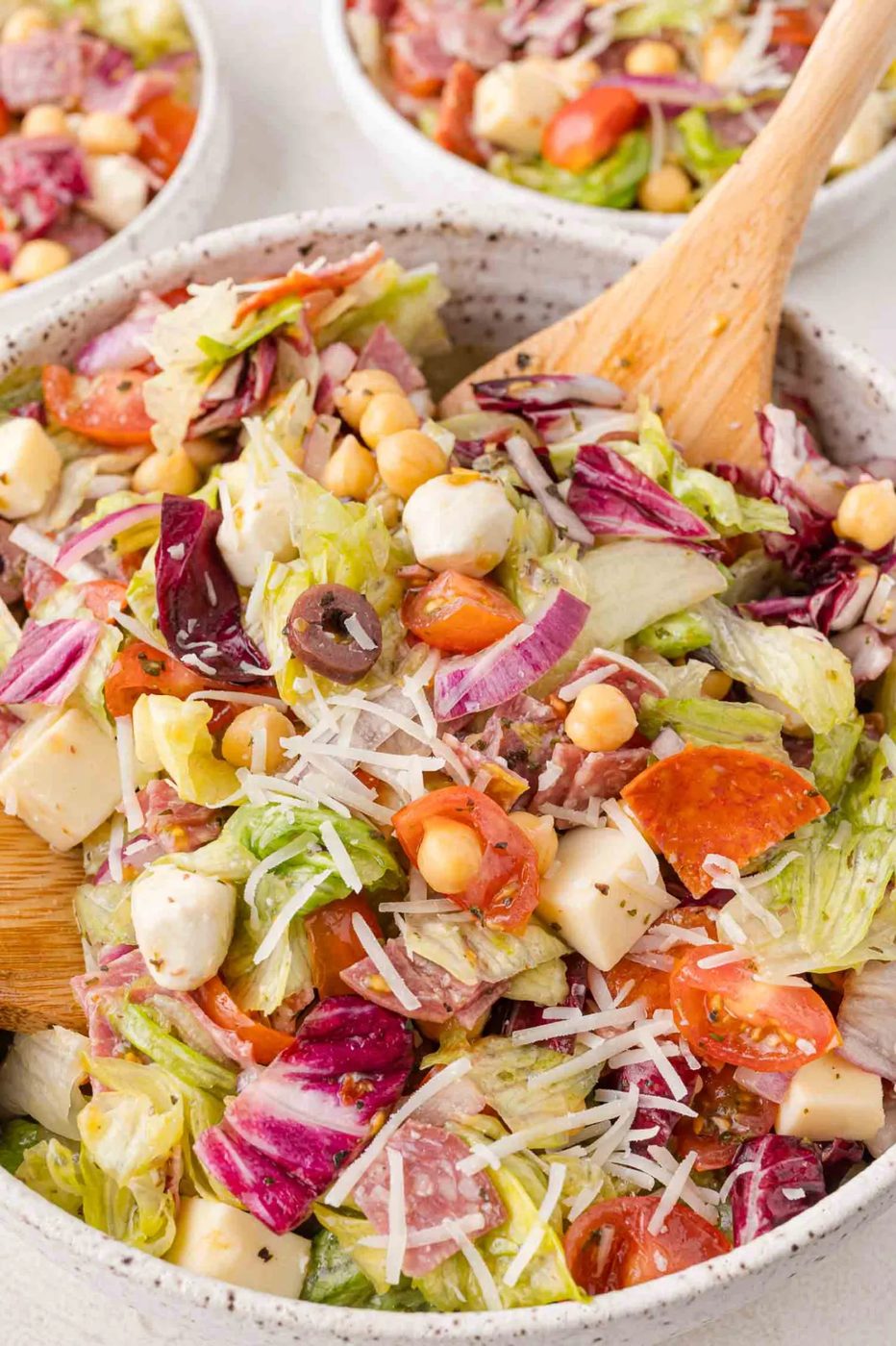 A mixed salad in a bowl featuring lettuce, tomatoes, cheese cubes, olives, chickpeas, and grated cheese, with a wooden spoon. Other bowls of salad are visible in the background.