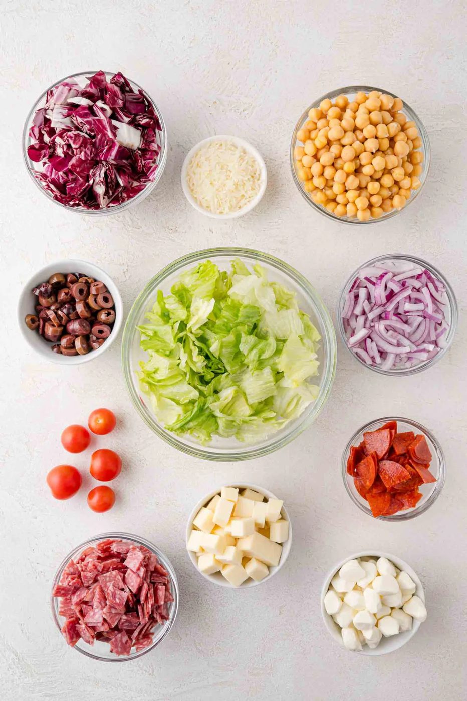 Bowls of salad ingredients including lettuce, chickpeas, radicchio, red onions, cheese, cherry tomatoes, olives, pepperoni, and salami, arranged on a white surface.