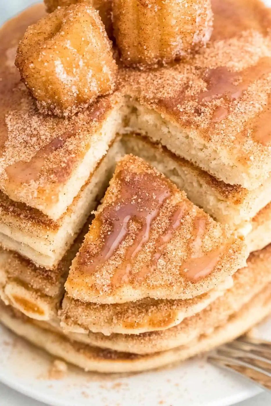 A stack of cinnamon-sugar pancakes with a wedge cut out, topped with churros and drizzled with syrup on a white plate.