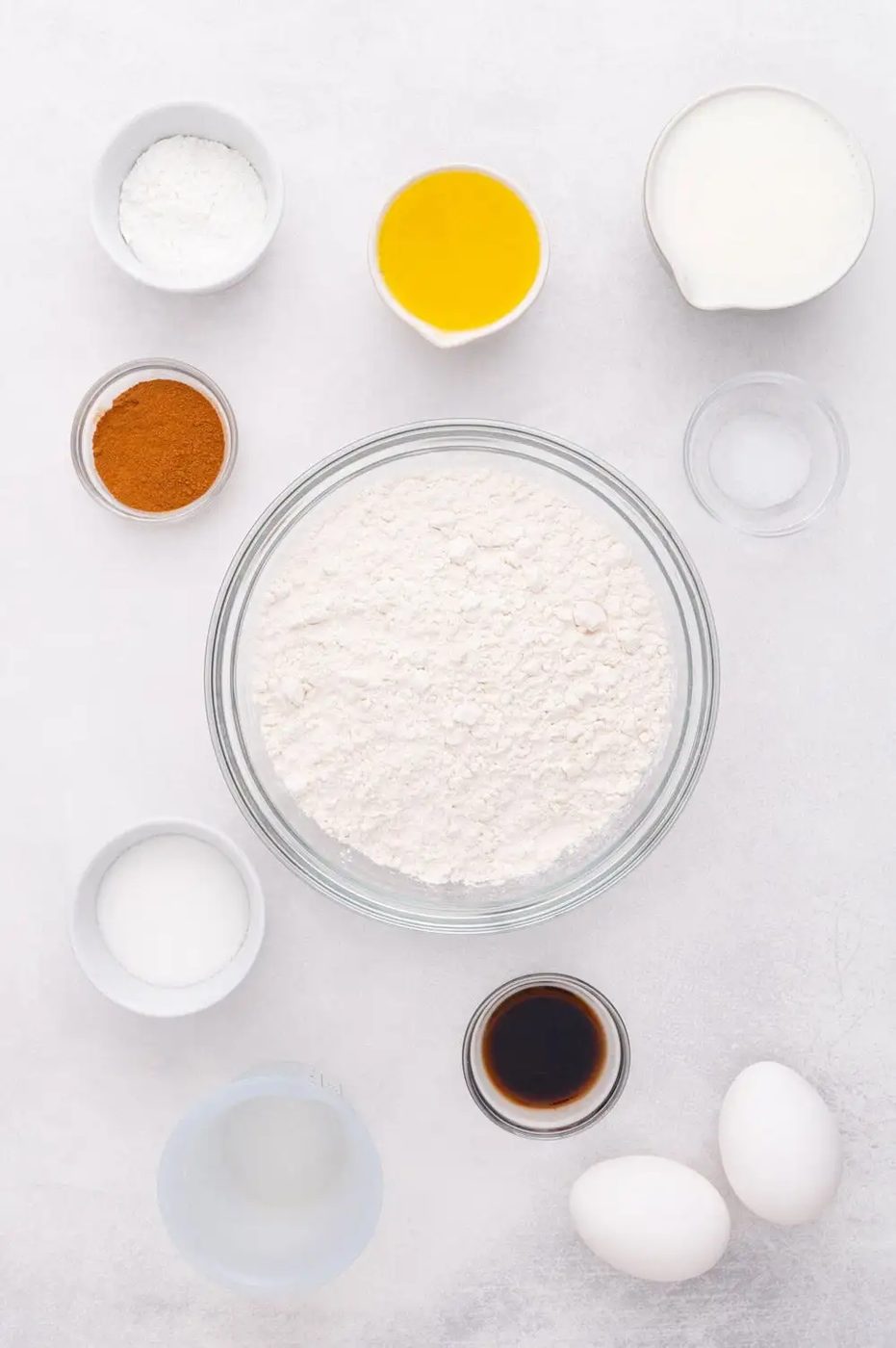 A variety of ingredients for baking are arranged on a white surface, including flour, eggs, vanilla, oil, sugar, cinnamon, baking powder, and milk.