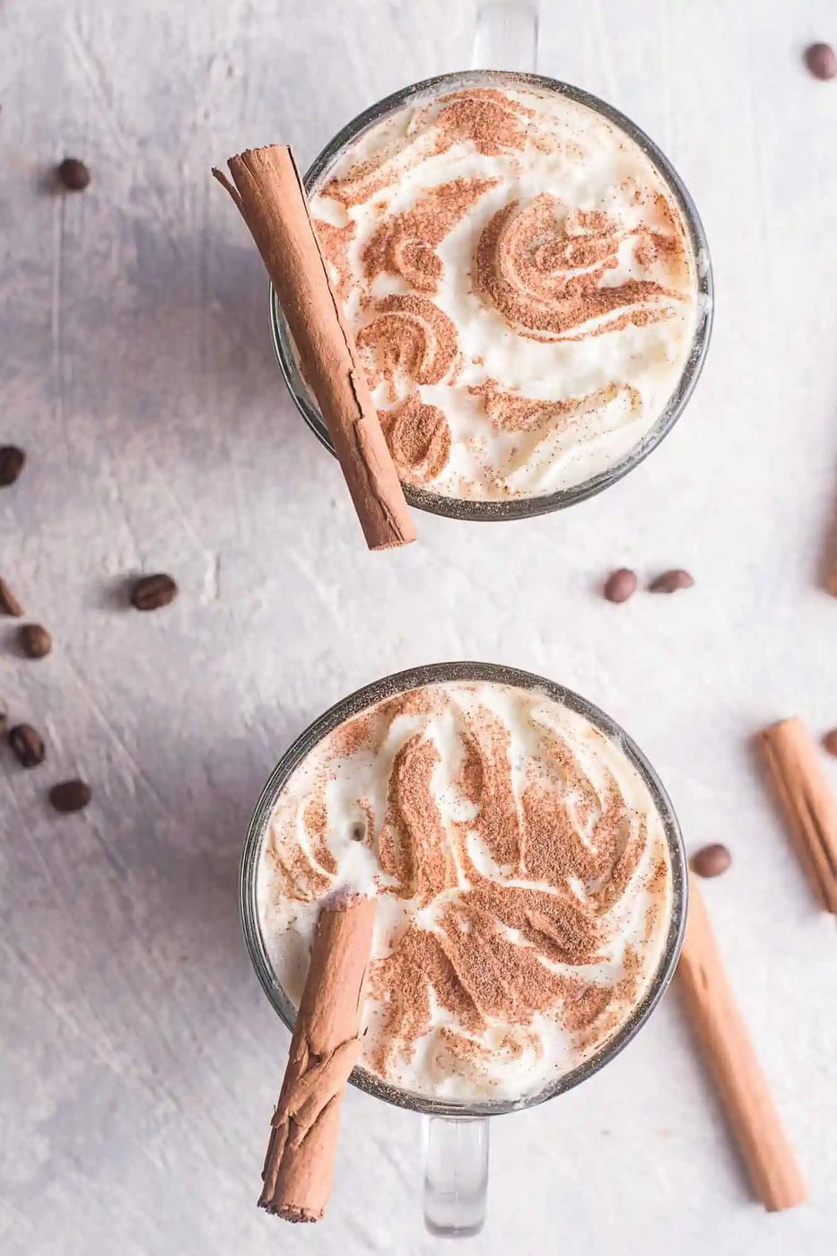 Two mugs of cinnamon dolce latte topped with cinnamon sticks and sprinkled cinnamon, surrounded by scattered coffee beans and cinnamon sticks.