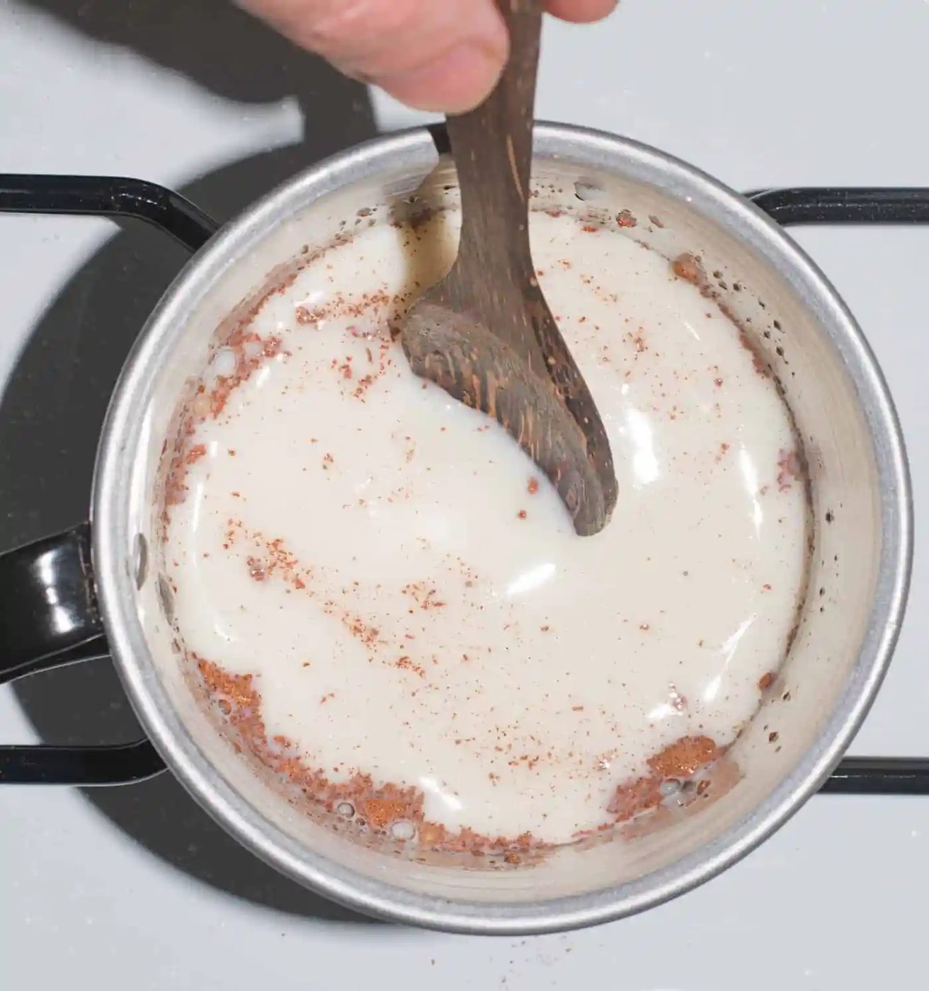 A wooden spoon stirs a mixture of milk and cocoa powder in a small metal pot on a stove.