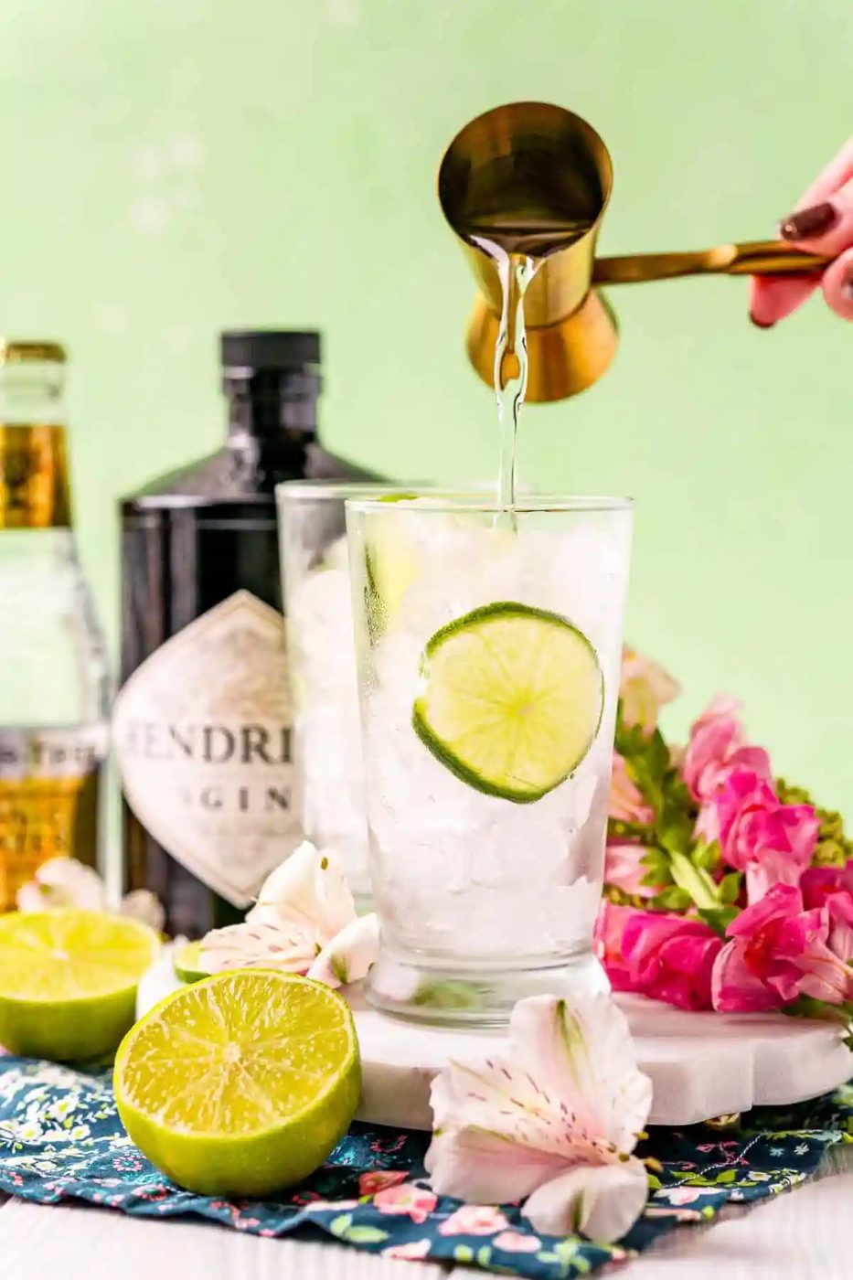 Gin being poured into a glass, a bottle of gin and a bottle of tonic pictured in the background.