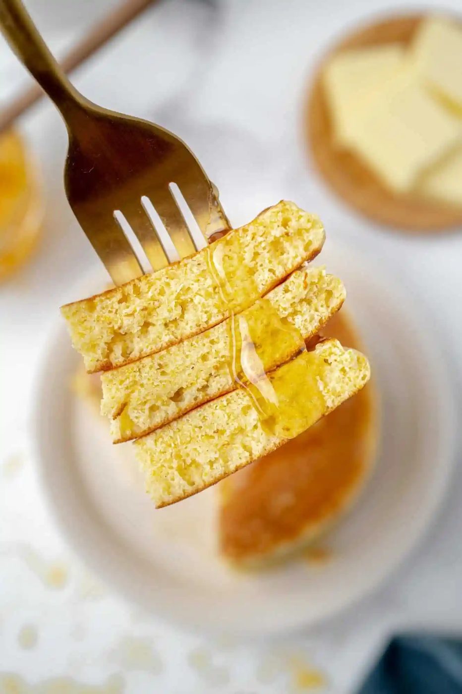 A fork holding three syrup-drizzled pancake pieces above a plate with pancakes and a butter dish in the background.
