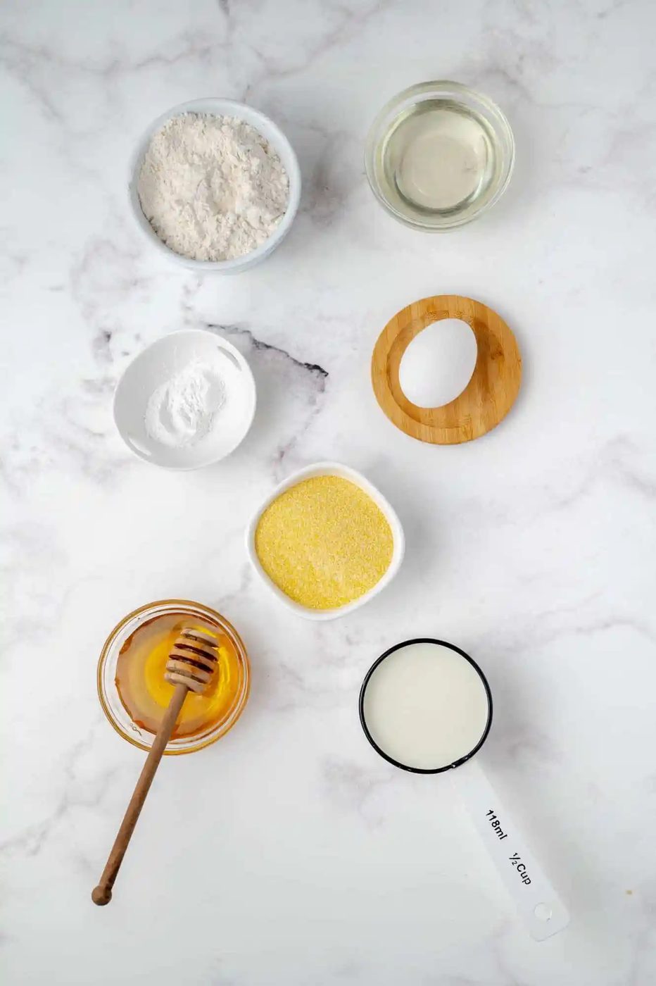 Ingredients on a marble surface: flour, oil, baking powder, cornmeal, honey with dipper, milk in a measuring cup, and an empty wooden lid.