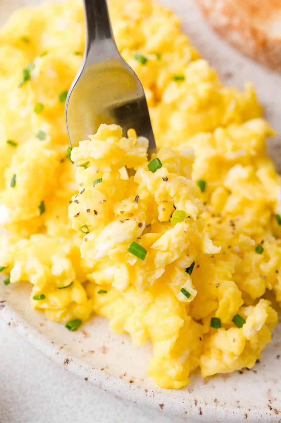 A fork holding a portion of scrambled eggs garnished with chopped chives and black pepper on a speckled plate.
