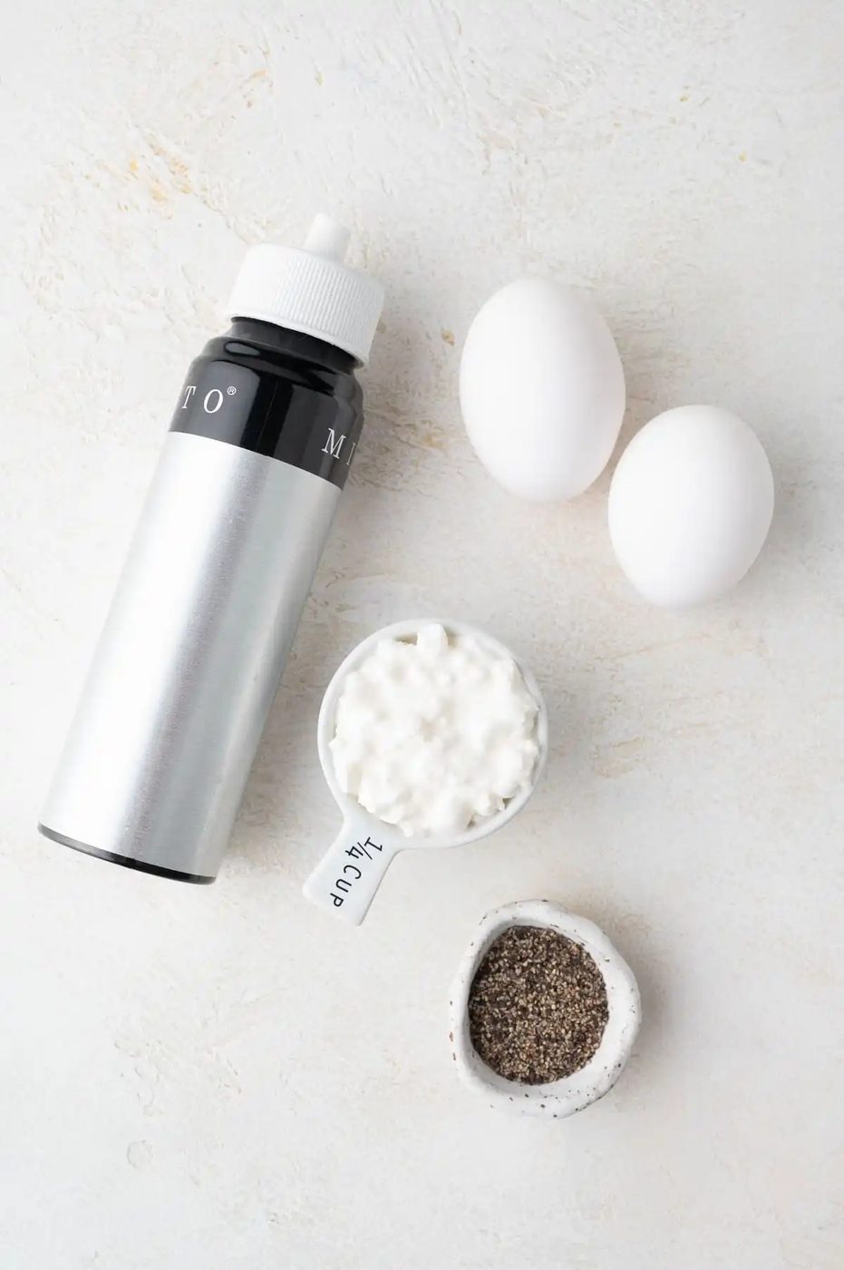 A bottle of oil spray, two eggs, a quarter cup of cottage cheese, and a small bowl of ground pepper are arranged on a light surface.