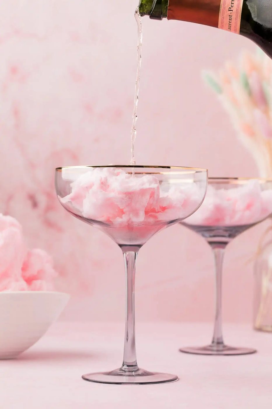 Champagne being poured into a glass filled with pink cotton candy, with another similar glass and a bowl of cotton candy in the background, set against a pink backdrop.