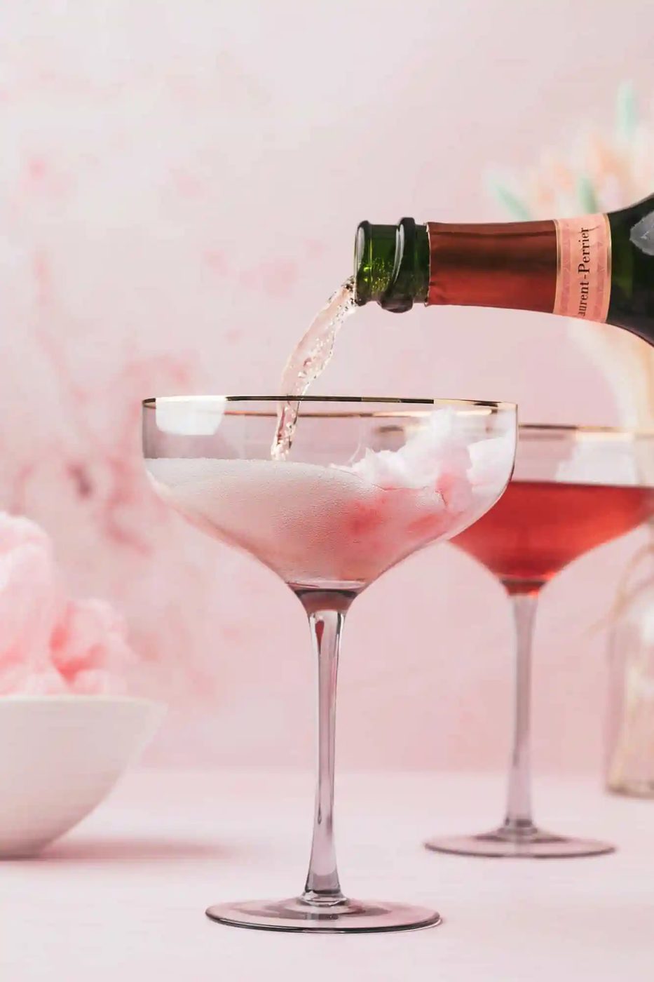 Champagne being poured into a glass with cotton candy inside, with another similar drink and a bowl of cotton candy in the background.