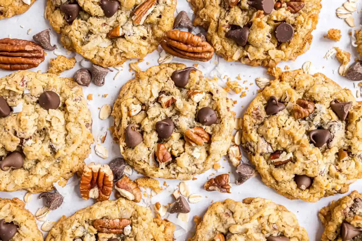 A close-up of oatmeal cookies topped with chocolate chips and pecans, with scattered pecans, oats, and chocolate pieces on a white surface.