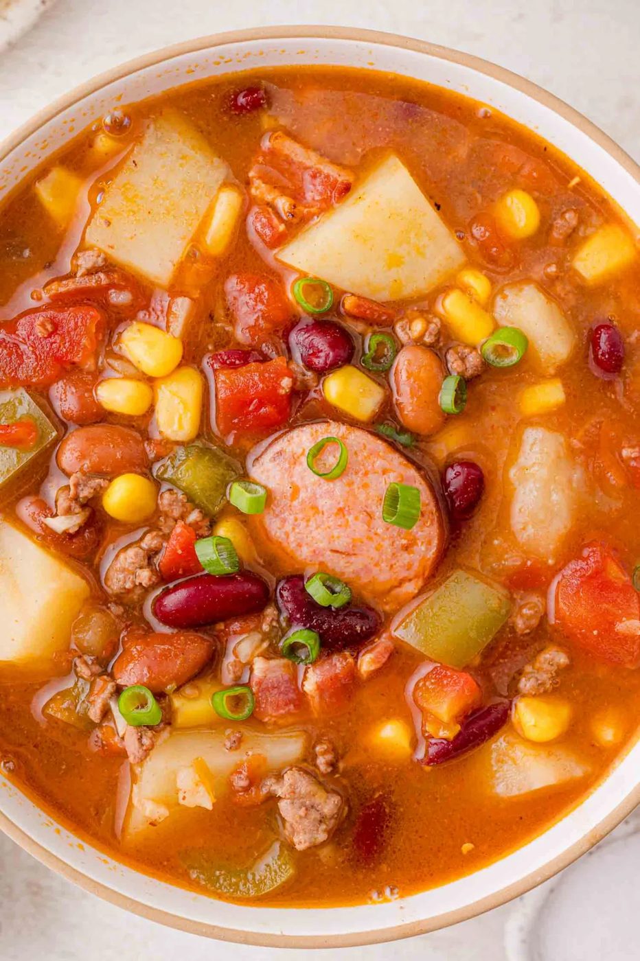 A bowl of hearty cowboy stew with sausage, potatoes, beans, corn, green peppers, and diced tomatoes, topped with sliced green onions.