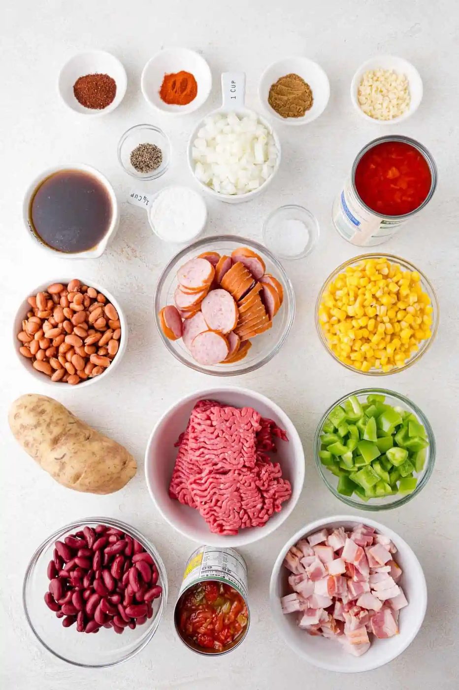 Assorted ingredients on a white surface, including beans, ground meat, bacon, corn, bell pepper, sausages, potato, canned tomatoes, and various seasonings in bowls.