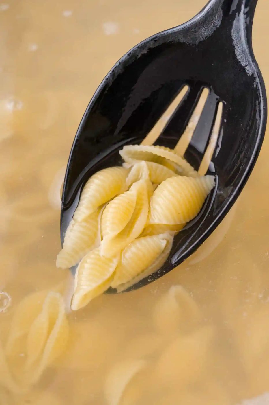 A black slotted spoon holding cooked shell pasta above a pot of boiling water.