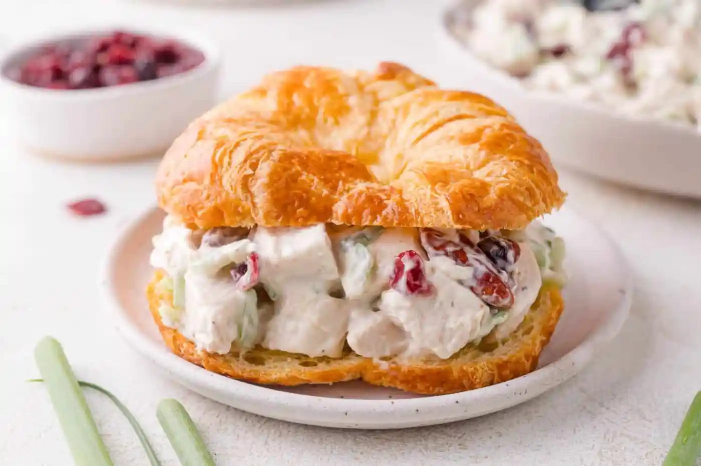 A croissant sandwich filled with chicken salad, diced celery, and dried cranberries on a white plate.