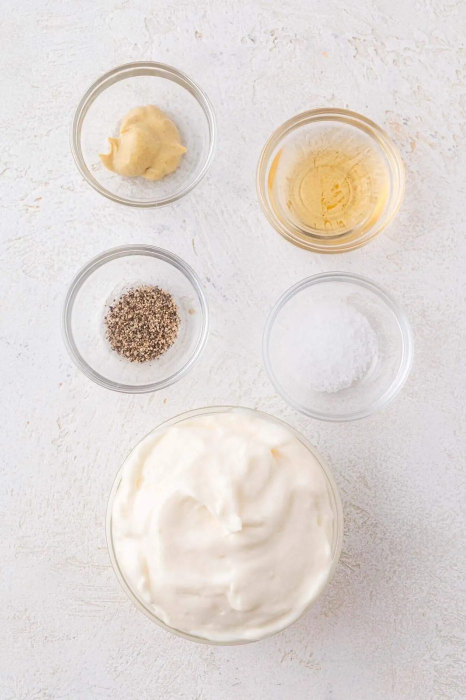 Five glass bowls containing mayonnaise, Dijon mustard, apple cider vinegar, ground black pepper, and salt on a light textured surface.