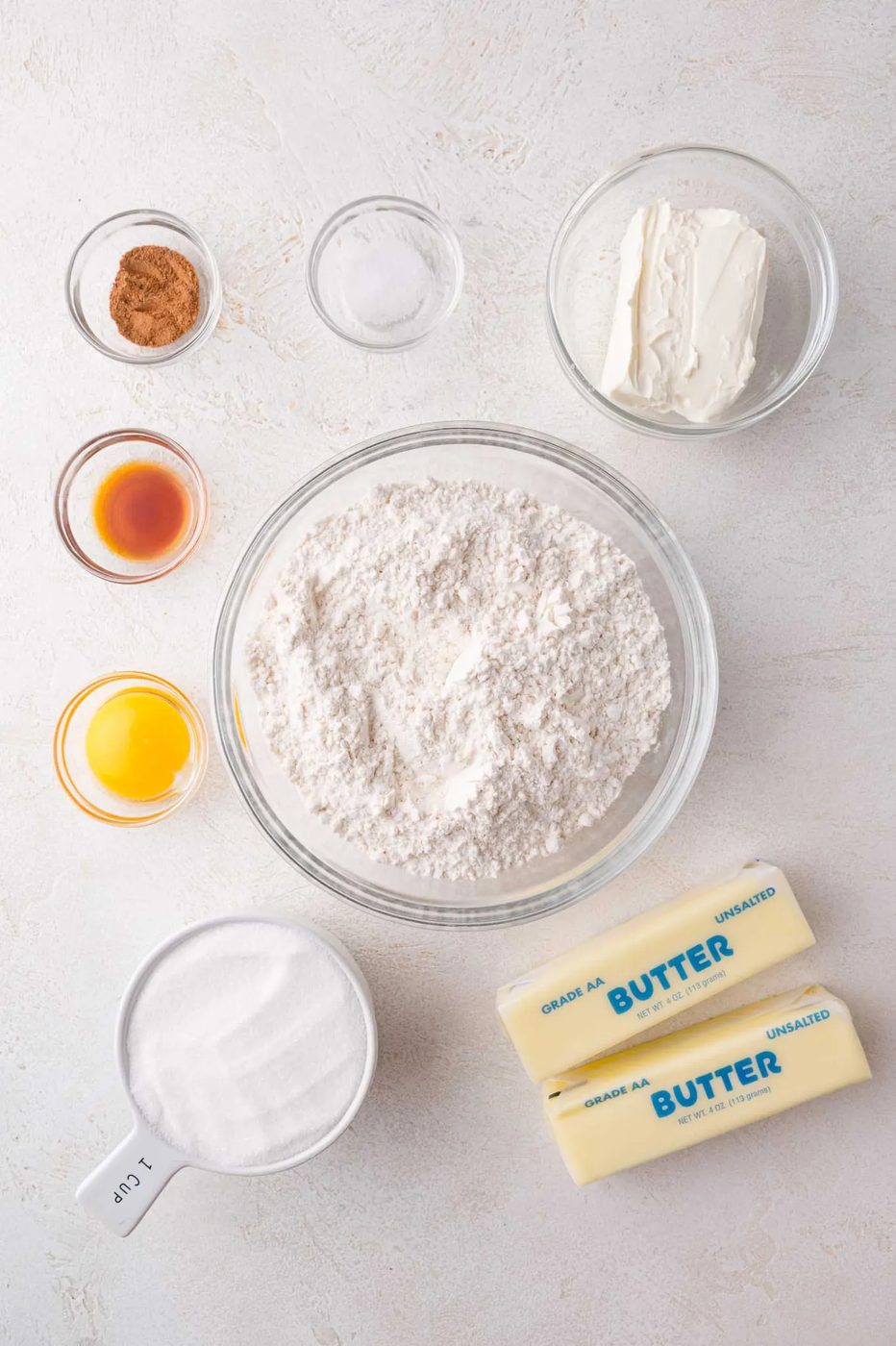 Various baking ingredients, including flour, two butter sticks, sugar, an egg yolk, vanilla, cream cheese, cinnamon, and salt, arranged in bowls and containers on a light surface.