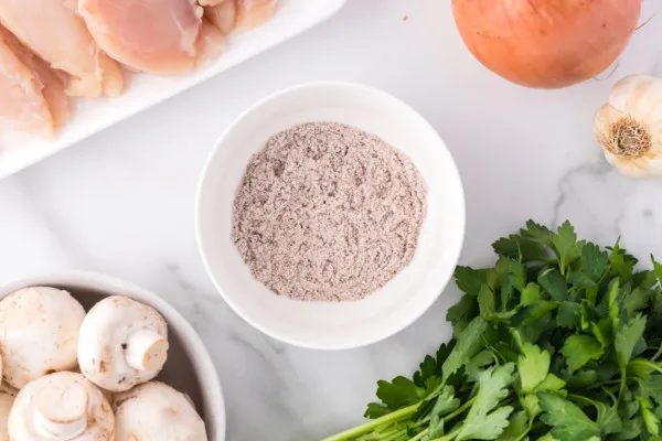 Overhead view of chicken seasoning in a bowl next to a bunch of parsley, a bowl of mushrooms, and a plate of uncooked chicken.