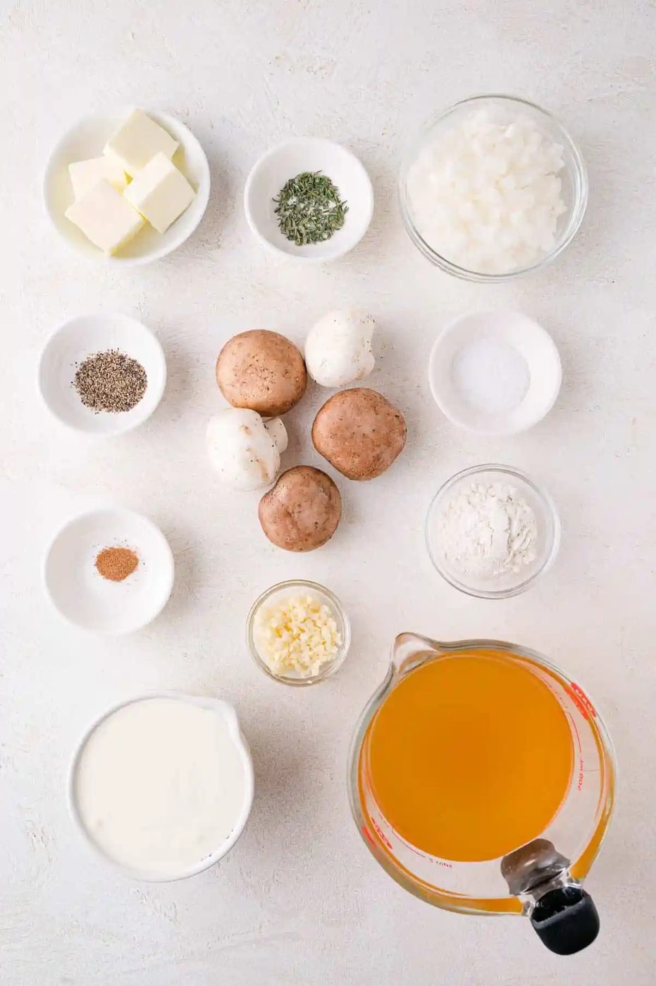 Ingredients arranged on a white surface include butter, onion, mushrooms, garlic, cream, broth, flour, black pepper, salt, a spice mix, and herbs.